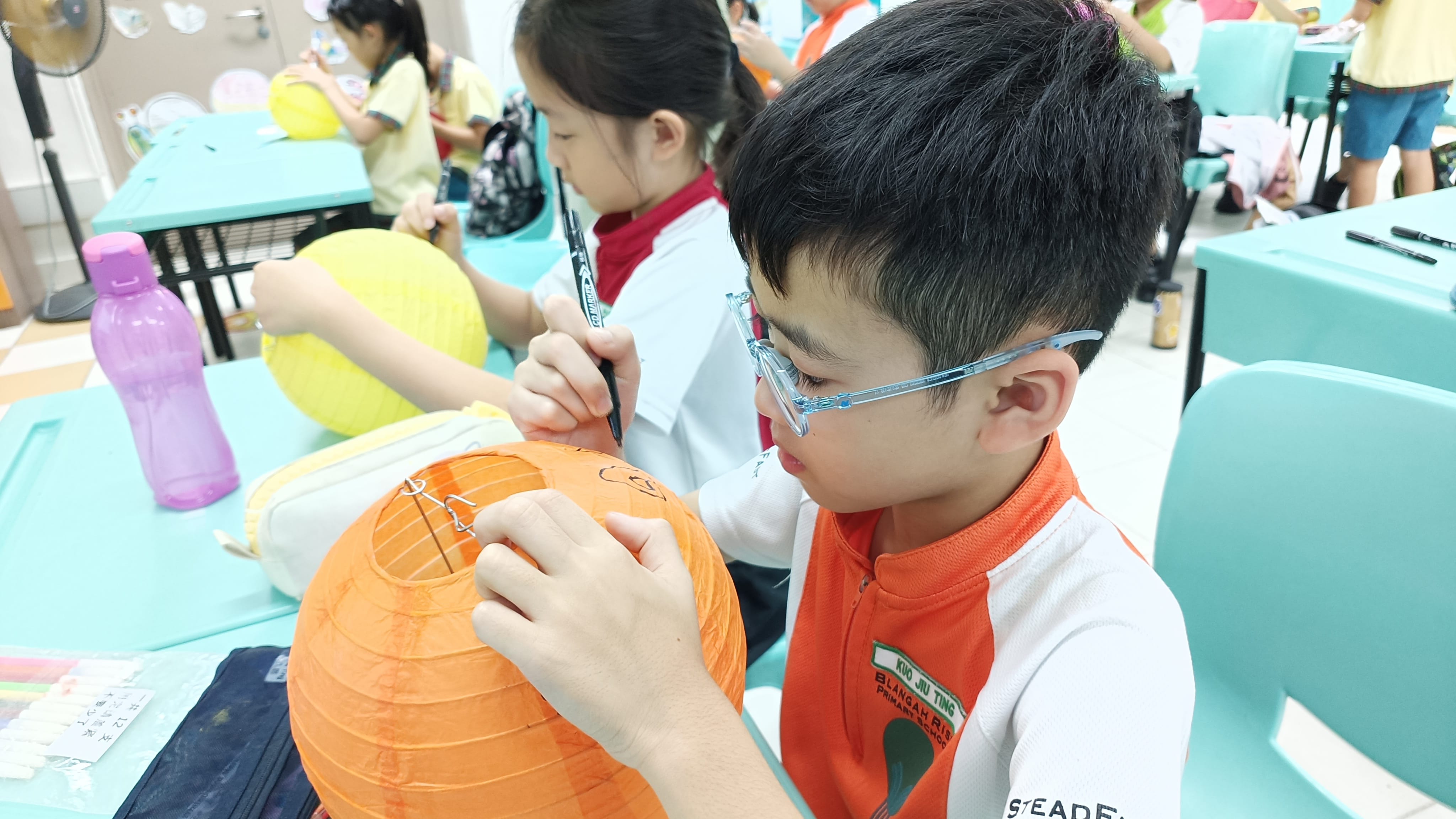 A young boy with glasses draws on an orange paper lantern while other children decorate lanterns nearby.